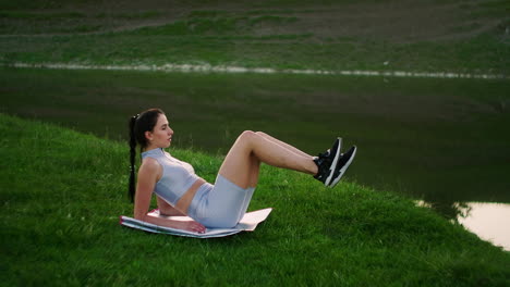 A-woman-does-exercises-for-the-muscles-of-the-press-lying-on-the-grass-in-a-Park-near-the-lake.-Work-with-abs-muscles.-Creating-a-beautiful-body